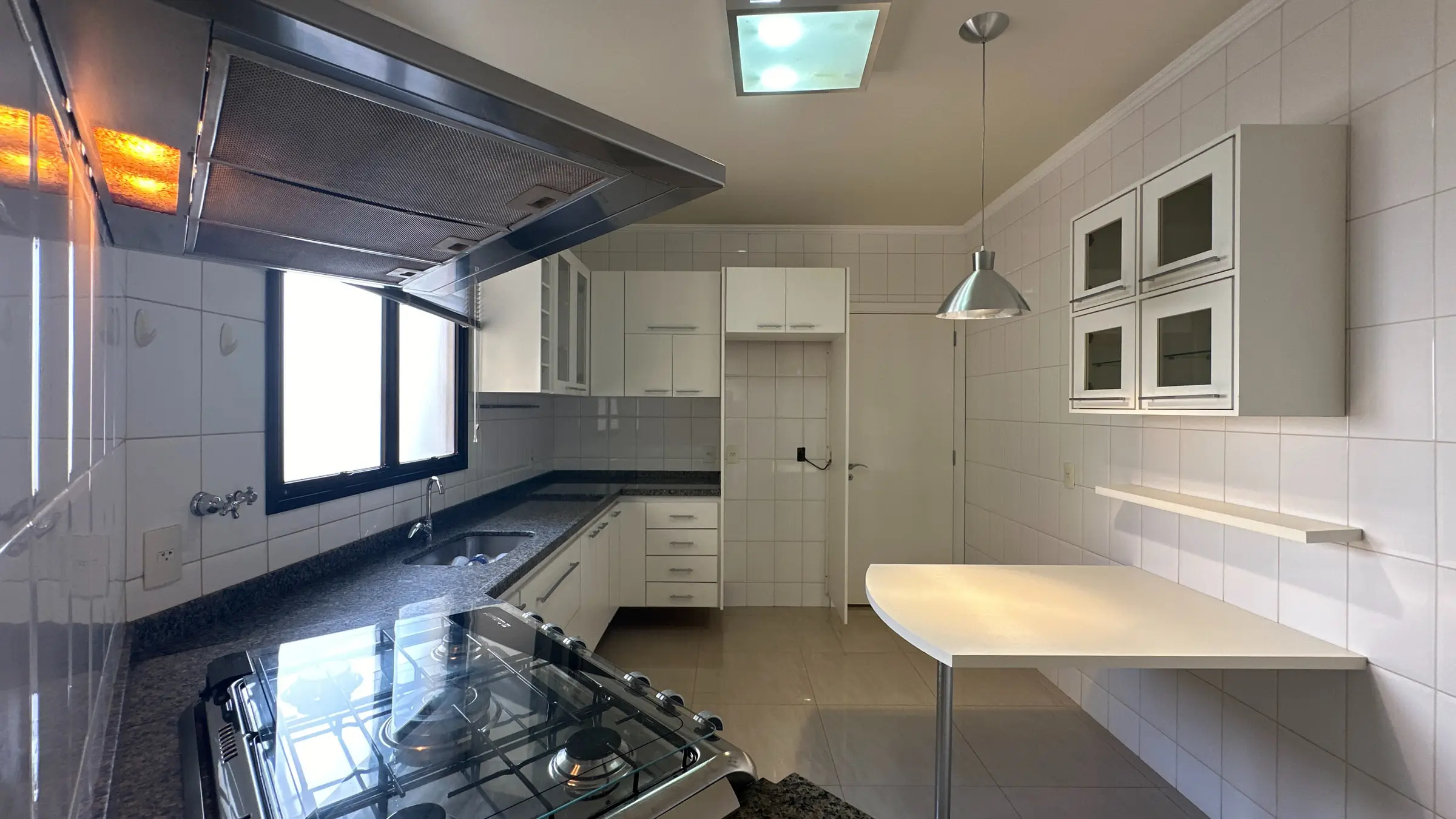 Outro ângulo da cozinha do apartamento, com coifa sobre o fogão e mesa de apoio
